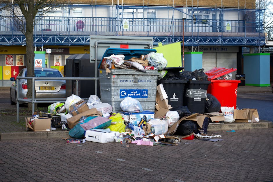A piled collection of black rubbish bags, some tied and others partially open, sits on the pavement in front of a red metal door attached to a rough-textured, off-white wall. The rubbish includes card